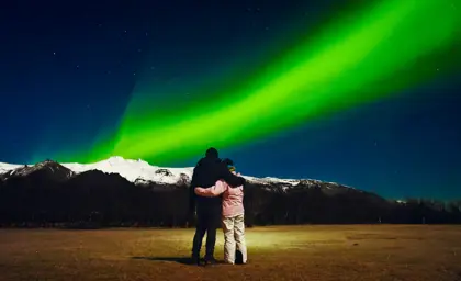 Couple embracing under bright green aurora borealis on a 3 day northern lights tour with Icelandic snowy mountains in the background at night.