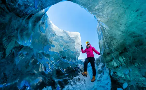 Explorer in a pink jacket climbing through sparkling blue ice formations inside a Jokulsarlon glacier cave on a guided adventure tour in Iceland.