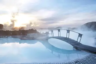Misty sunrise at Blue Lagoon Iceland with steam rising over the geothermal spa and a wooden bridge reflecting in the tranquil blue waters.