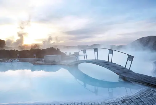 Misty sunrise at Blue Lagoon Iceland with steam rising over the geothermal spa and a wooden bridge reflecting in the tranquil blue waters.