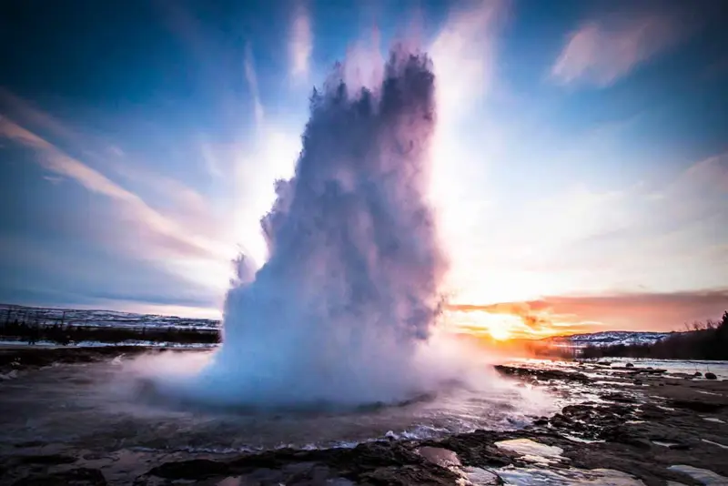 Strokkur Geyser erupting at sunrise in Iceland, showcasing a dramatic water plume against a colorful sky with a snowy winter landscape in the background.