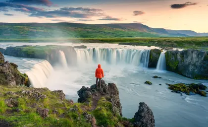Male tourist in orange jacket standing on rocky cliffs overlooking Godafoss waterfall during a vibrant Icelandic sunset