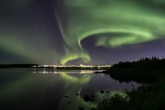 Mesmerizing view of the Northern Lights dancing over Reykjavik, reflecting in the serene waters of a lagoon at night.