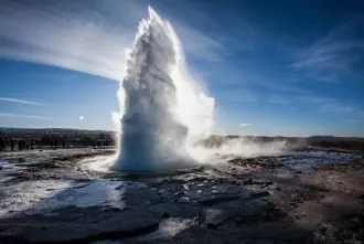 Powerful eruption of a geyser under a clear blue sky during a Golden Circle tour in Iceland, surrounded by icy terrain and curious onlookers.