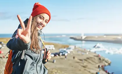Young woman in a red knitted hat smiling and flashing a peace sign at Jökulsárlón Glacier Lagoon in Iceland with icebergs and bridge in the background.