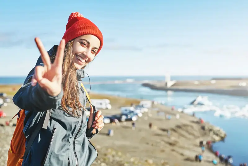 Young woman in a red knitted hat smiling and flashing a peace sign at Jökulsárlón Glacier Lagoon in Iceland with icebergs and bridge in the background.