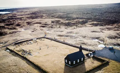 Aerial view of Búðakirkja, the iconic black church in Iceland, surrounded by a stone wall and a rugged volcanic landscape, with the Atlantic Ocean visible in the background.