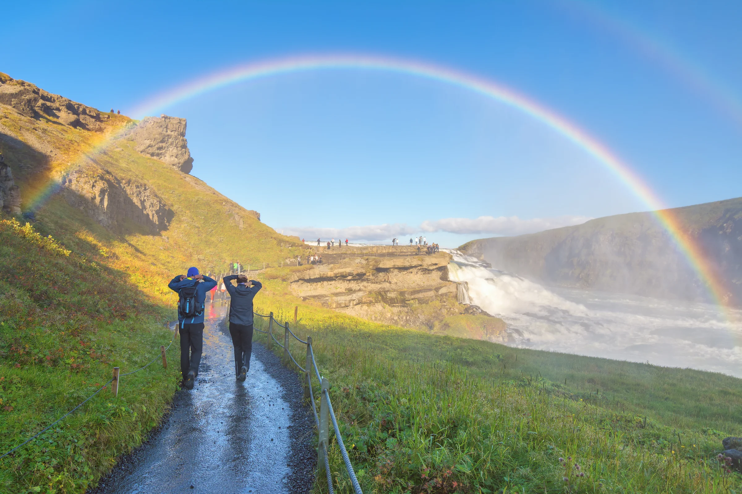 A couple on Iceland's 5 Day Ring Road Tour walking towards the stunning Gullfoss waterfall in Iceland beneath a bright rainbow.
