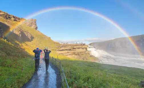 A couple on Iceland's 5 Day Ring Road Tour walking towards the stunning Gullfoss waterfall in Iceland beneath a bright rainbow.