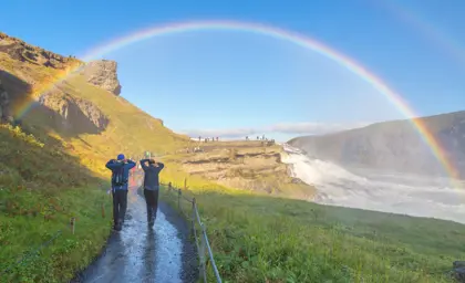 A couple on Iceland's 5 Day Ring Road Tour walking towards the stunning Gullfoss waterfall in Iceland beneath a bright rainbow.