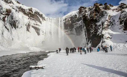 Visitors admiring the icy Skógafoss waterfall with a rainbow in the mist, during a winter tour in Iceland.