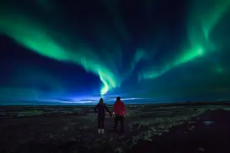Couple on a 5 day northern lights tour holding hands watching vivid green northern lights dance over Icelandic plains under a deep midnight sky .
