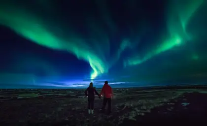 Couple on a 5 day northern lights tour holding hands watching vivid green northern lights dance over Icelandic plains under a deep midnight sky .