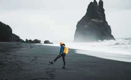 Young woman with yellow backpack and hat walks across Reynisfjara black sand beach toward dramatic rock formations in South Iceland.