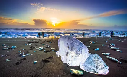 Stunning sunset over Diamond Beach in Iceland with large transparent ice chunks scattered across the black sand shore and silhouettes of tourists in the distance.