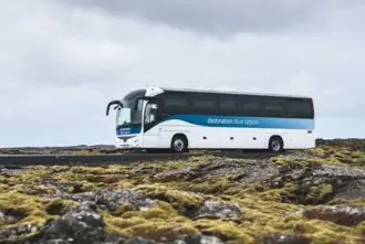 A Destination Blue Lagoon bus driving on a road surrounded by a moss covered lava field.