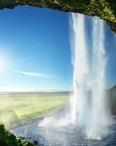 Majestic summer view from behind Seljalandsfoss Waterfall, on Iceland's five day guided tour, with bright sunlight illuminating the green landscape.