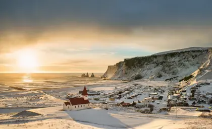 Sunset over the snow-covered village of Vík í Mýrdal in Iceland, with the iconic red-roofed church in the foreground and the Reynisdrangar sea stacks rising from the ocean in the distance