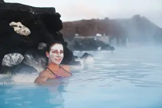 Smiling woman enjoying a silica face mask while relaxing in the milky-blue waters of Iceland’s Blue Lagoon, surrounded by volcanic rocks – a rejuvenating Travel Reykjavik experience.