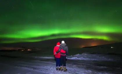 A couple in winter clothing embracing while watching the Northern Lights in Iceland, with a vibrant green aurora illuminating the night sky.