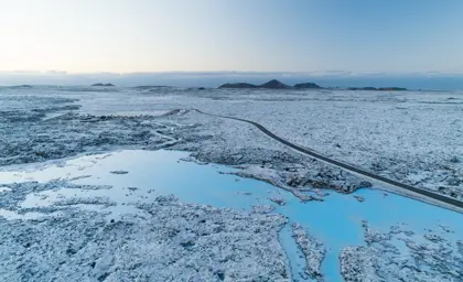 Winter aerial view of the Blue Lagoon geothermal spa, surrounded by volcanic lava fields and snow, showcasing the stunning icy blue water and a winding road leading to the spa in Iceland.