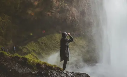 Adventurer in a raincoat standing behind Seljalandsfoss waterfall, surrounded by mist and mossy cliffs on a scenic Icelandic nature tour.