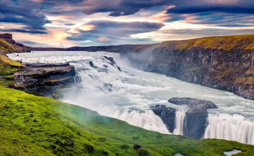 Scenic view of Gullfoss Waterfall during a vibrant sunset, with tourists walking along the green path as part of the Golden Circle tour in Iceland.