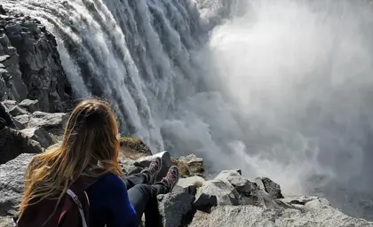 Woman sitting on rocky edge overlooking the roaring cascade of Dettifoss waterfall in Iceland.