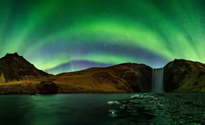 Northern Lights dancing above Skógafoss waterfall in Iceland, with vivid green auroras illuminating the night sky and reflecting on the waters below, creating a stunning natural spectacle.