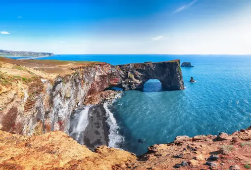 Scenic view of Dyrhólaey rock arch, a natural coastal landmark in South Iceland, with clear blue skies and ocean waves crashing on the black sand beach below.