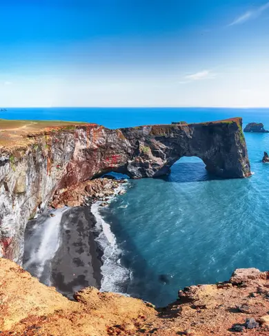 Scenic view of Dyrhólaey rock arch, a natural coastal landmark in South Iceland, with clear blue skies and ocean waves crashing on the black sand beach below.