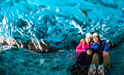 Couple dressed in winter gear sitting inside a vibrant blue ice cave in Jökulsárlón, Iceland, during a guided tour experience.