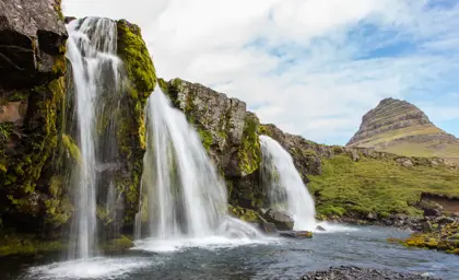Majestic Kirkjufellsfoss waterfall with the iconic Kirkjufell mountain in the background, surrounded by lush greenery and flowing water, a must-see stop on Iceland's Snæfellsnes Peninsula tours.