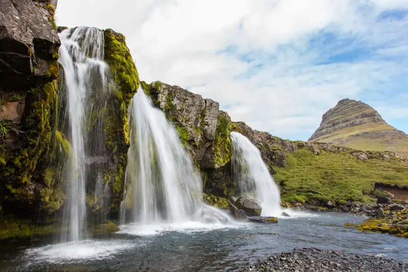 Majestic Kirkjufellsfoss waterfall with the iconic Kirkjufell mountain in the background, surrounded by lush greenery and flowing water, a must-see stop on Iceland's Snæfellsnes Peninsula tours.