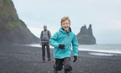 Smiling boy in blue jacket runs along Reynisfjara black sand beach with sea stacks and father in the background in South Iceland.