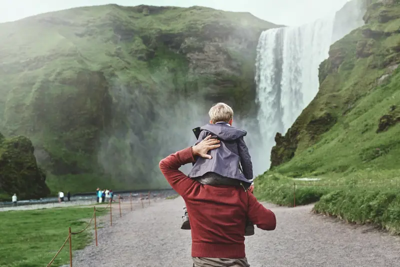 Father carrying son on shoulders while walking towards the misty Skógafoss waterfall in southern Iceland on a family trip.