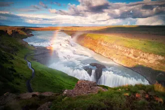 Gullfoss waterfall with a vibrant rainbow arcing over the cascading water, surrounded by lush green landscape and a scenic pathway on Iceland’s Golden Circle.
