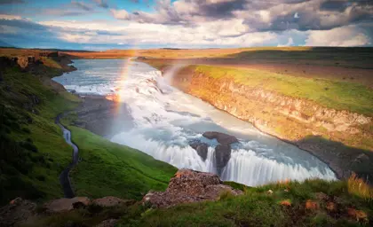 Gullfoss waterfall with a vibrant rainbow arcing over the cascading water, surrounded by lush green landscape and a scenic pathway on Iceland’s Golden Circle.