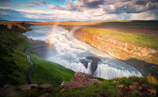 Gullfoss waterfall with a vibrant rainbow arcing over the cascading water, surrounded by lush green landscape and a scenic pathway on Iceland’s Golden Circle.