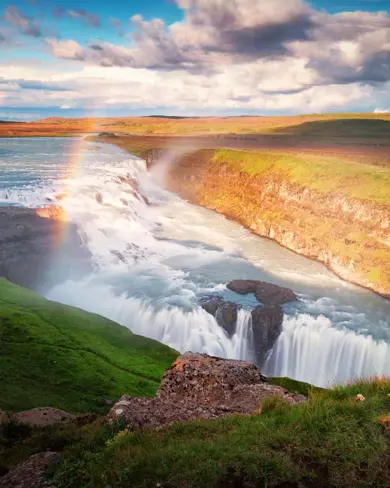 Gullfoss waterfall with a vibrant rainbow arcing over the cascading water, surrounded by lush green landscape and a scenic pathway on Iceland’s Golden Circle.