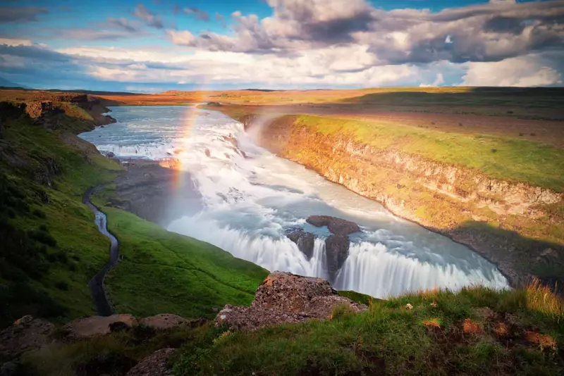 Gullfoss waterfall with a vibrant rainbow arcing over the cascading water, surrounded by lush green landscape and a scenic pathway on Iceland’s Golden Circle.