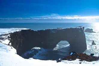 A stunning winter view of Dyrhólaey's natural arch and the Atlantic Ocean from a snow-covered cliff in Iceland, capturing the winter beauty of the South Coast.