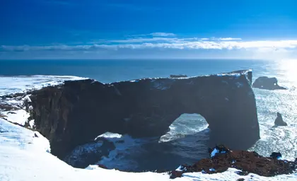 A stunning winter view of Dyrhólaey's natural arch and the Atlantic Ocean from a snow-covered cliff in Iceland, capturing the winter beauty of the South Coast.