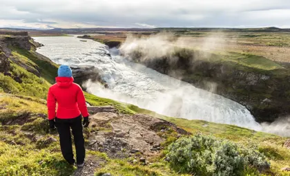 Woman in red jacket overlooking misty Gullfoss waterfall canyon in Iceland on a cloudy day