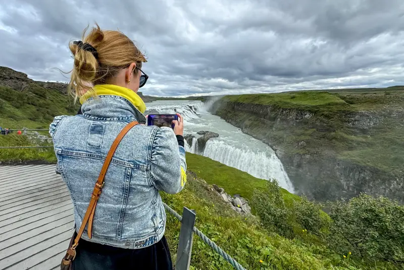 Tourist woman capturing the powerful Gullfoss waterfall in Iceland on a cloudy summer day from the viewing platform.