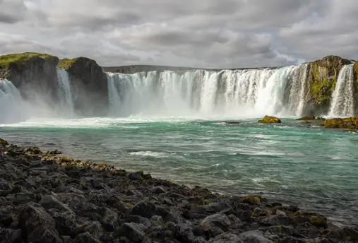 Scenic view of Goðafoss waterfall in Iceland with powerful turquoise waters cascading over rugged cliffs, framed by a rocky shoreline under a cloudy sky.