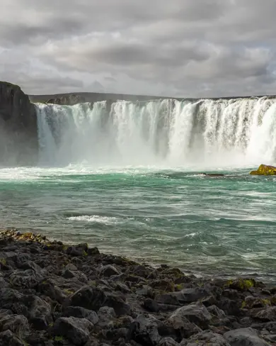 Scenic view of Goðafoss waterfall in Iceland with powerful turquoise waters cascading over rugged cliffs, framed by a rocky shoreline under a cloudy sky.