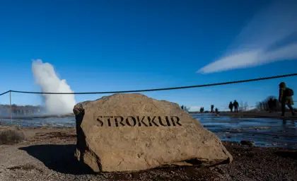 Strokkur geyser erupting in the background of a close up of rock marked with the geysers name.