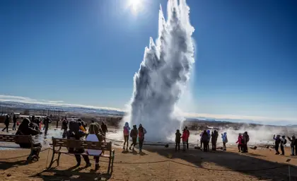 A crowd of tourists gathers to witness the powerful eruption of the Geysir hot spring in Iceland, surrounded by bright blue skies and the warm sun illuminating the landscape. Perfect for showcasing Icelandic natural wonders and outdoor activities.