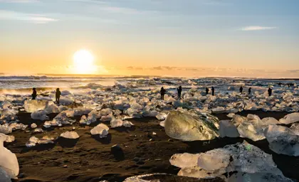 Tourists walking among glistening icebergs on black sand at Diamond Beach in Iceland during golden hour sunset.